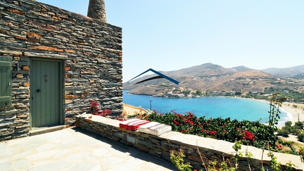 A stone building with a green door, a stone chimney, and a view of a blue sea and hills in the background.