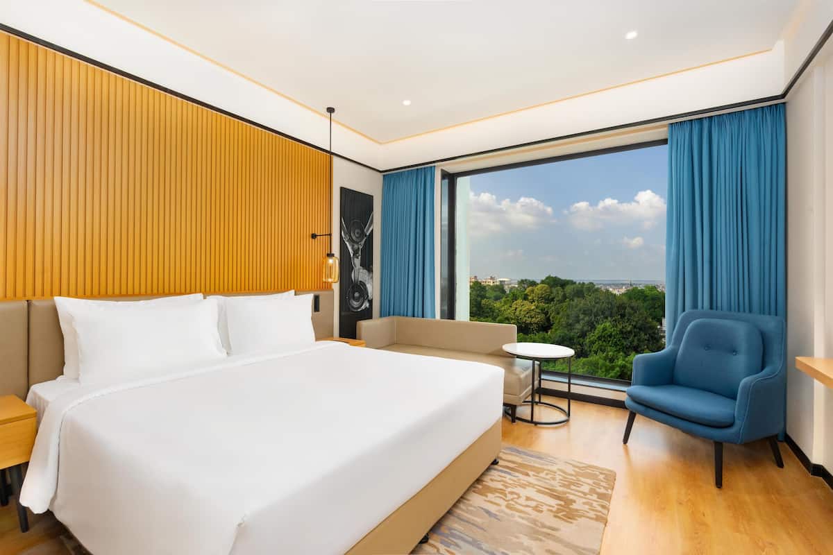 A modern hotel room with a large bed, a sofa, a blue armchair, and a view of the city through large windows.