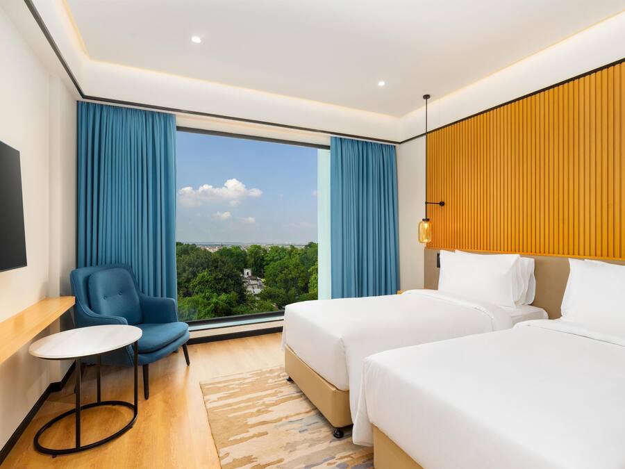 A hotel room with a large window, two beds, a chair, a small table, and a TV.