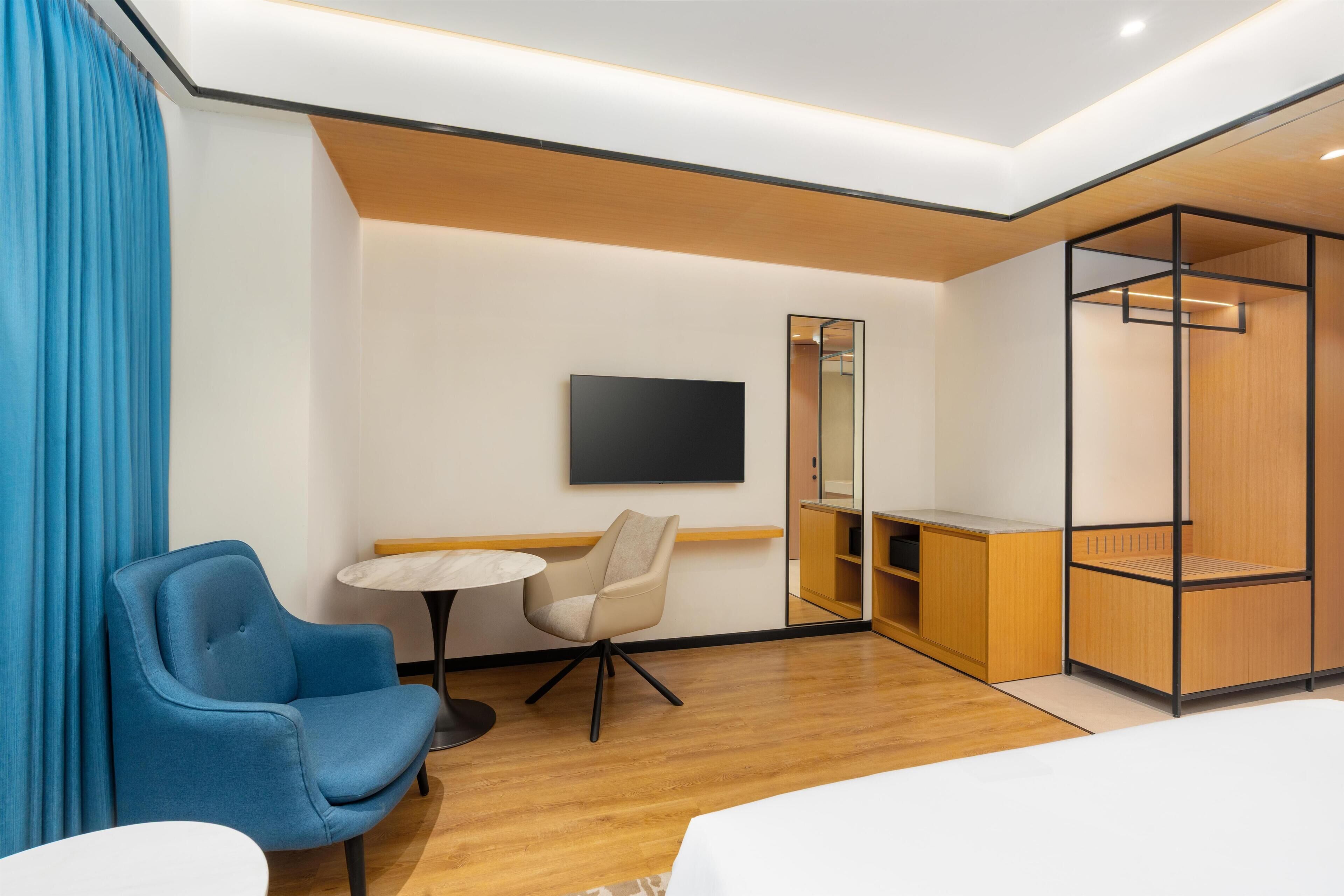 A hotel room with a bed, a blue armchair, a small round table, a TV mounted on the wall, and a wooden wardrobe.