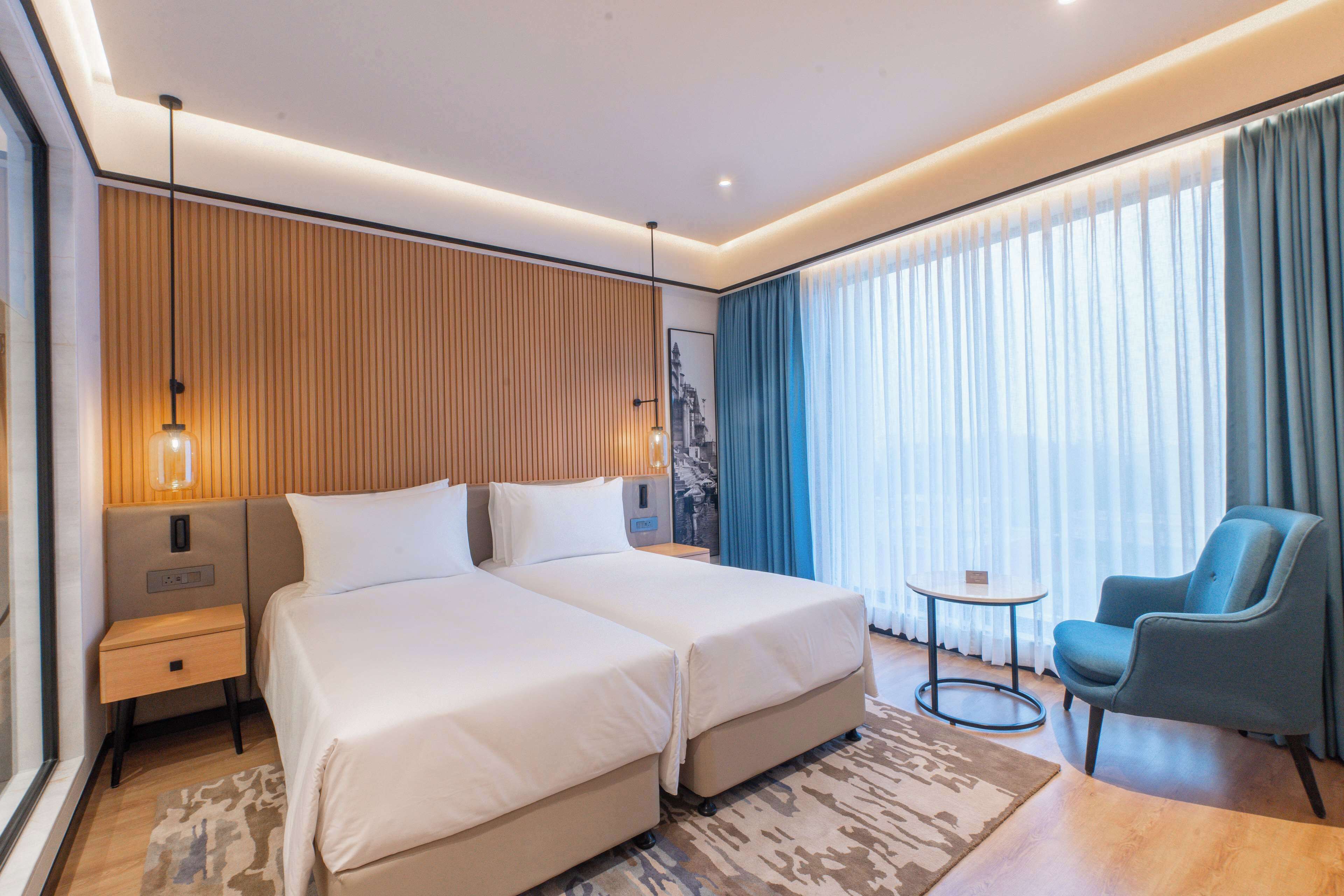A modern hotel room with two beds, a small round table, a blue armchair, and a large window with curtains.