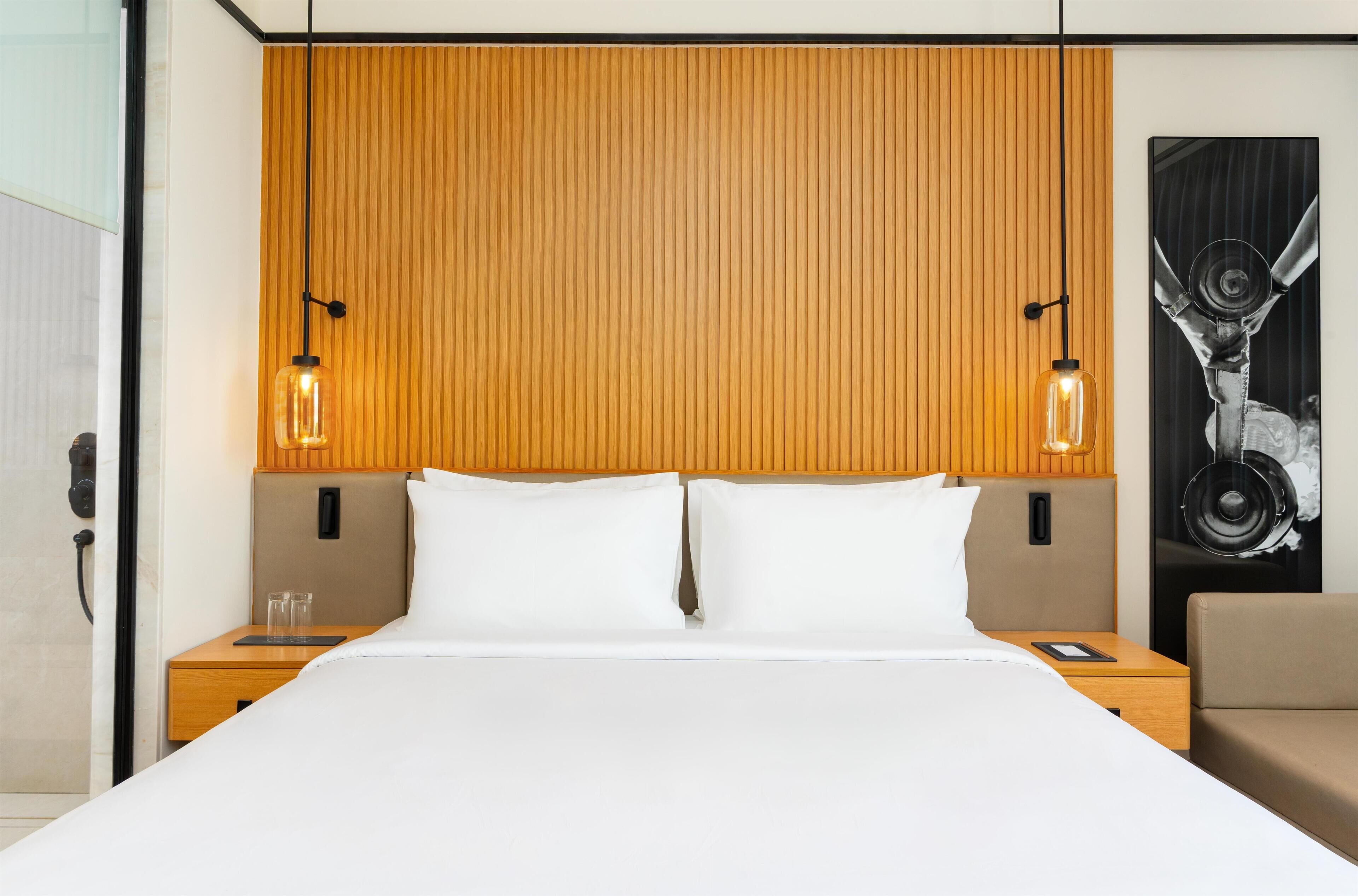 A hotel room with a wooden headboard, two bedside tables with lamps, a bed with white linens, and a large abstract painting on the wall.