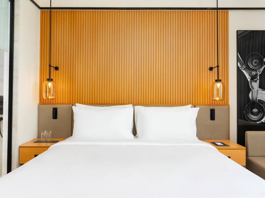 A hotel room with a wooden headboard, two bedside tables with lamps, a bed with white linens, and a large abstract painting on the wall.