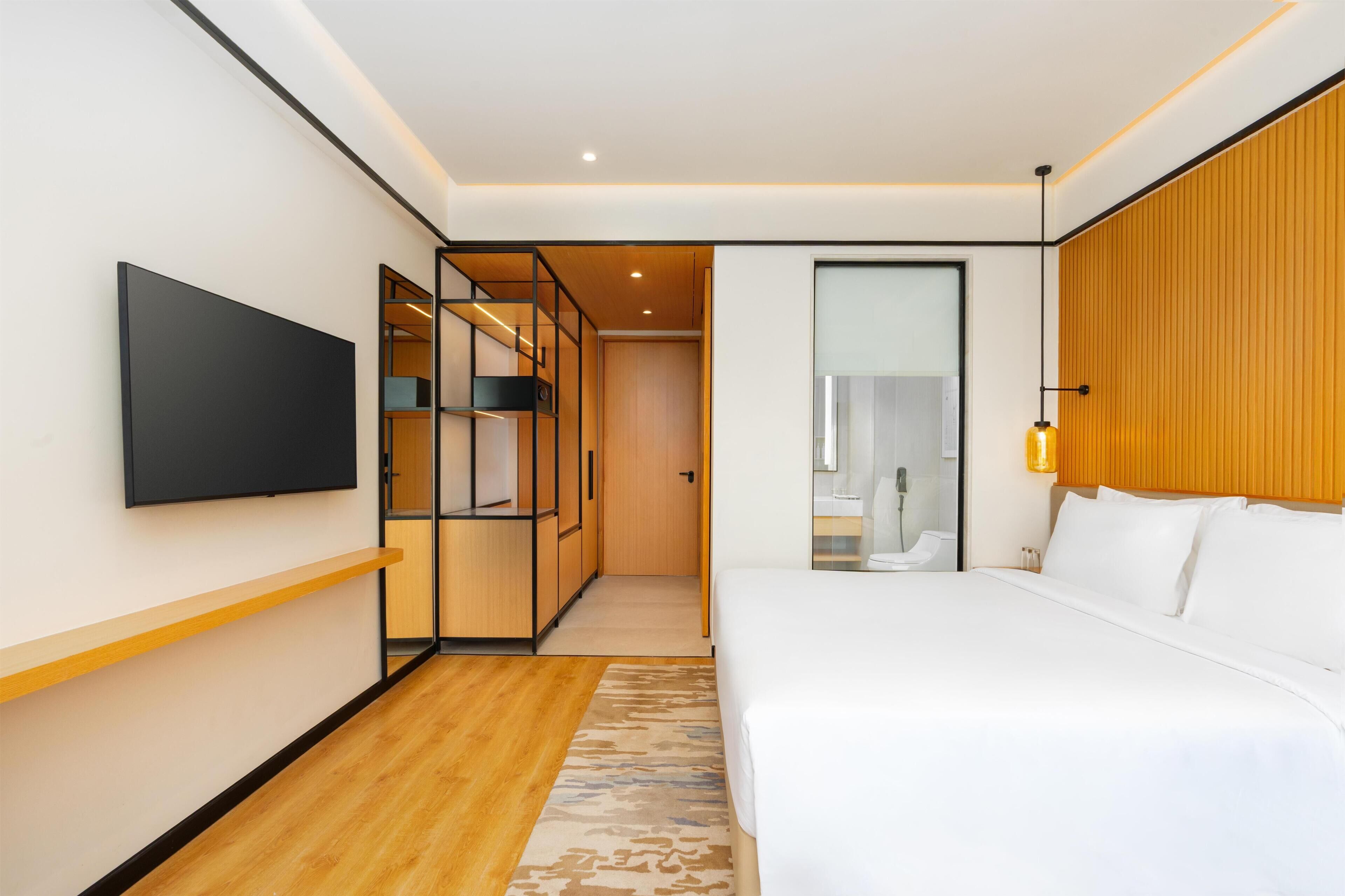 A modern hotel room with a large bed, a flat-screen TV mounted on the wall, a wooden wardrobe, and a bathroom visible through a glass door.