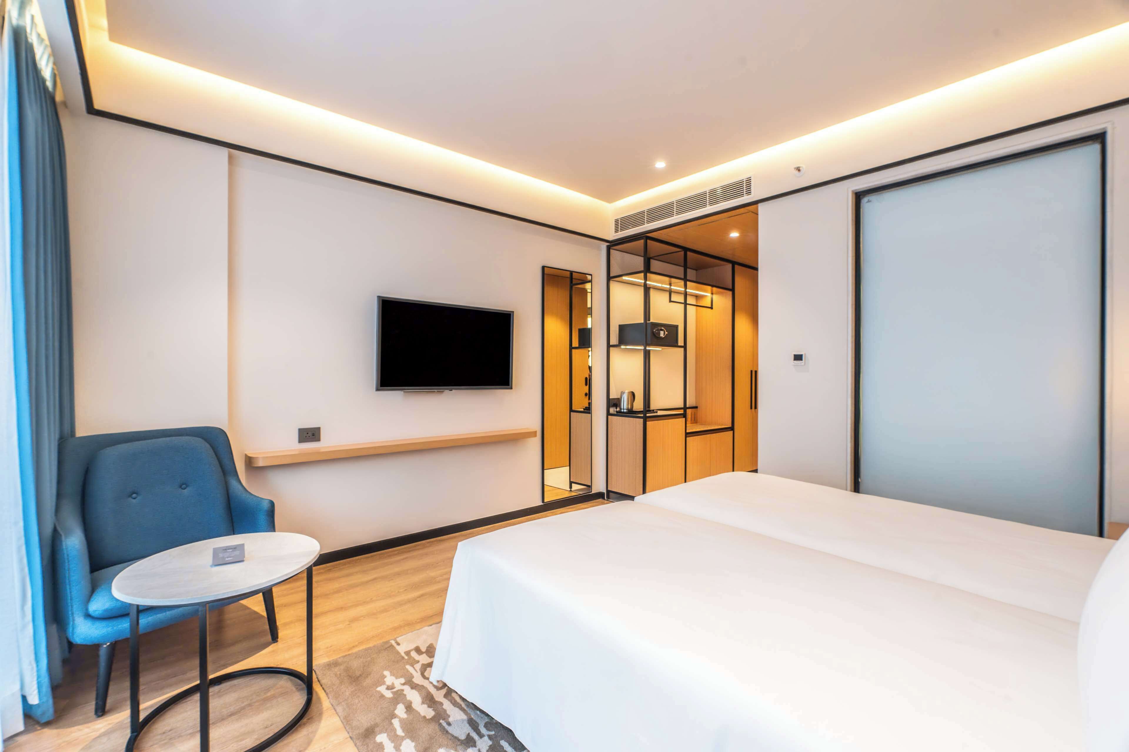A modern hotel room with a bed, a TV mounted on the wall, a seating area with a chair, and a small table.