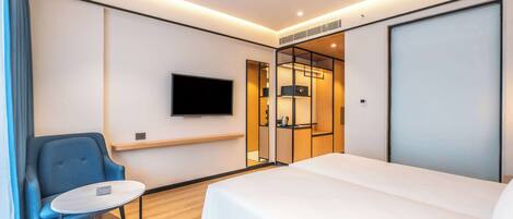 A modern hotel room with a bed, a TV mounted on the wall, a seating area with a chair, and a small table.