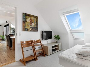 A bedroom with a sloped ceiling, a bed, a TV, a desk with chairs, and a painting on the wall.