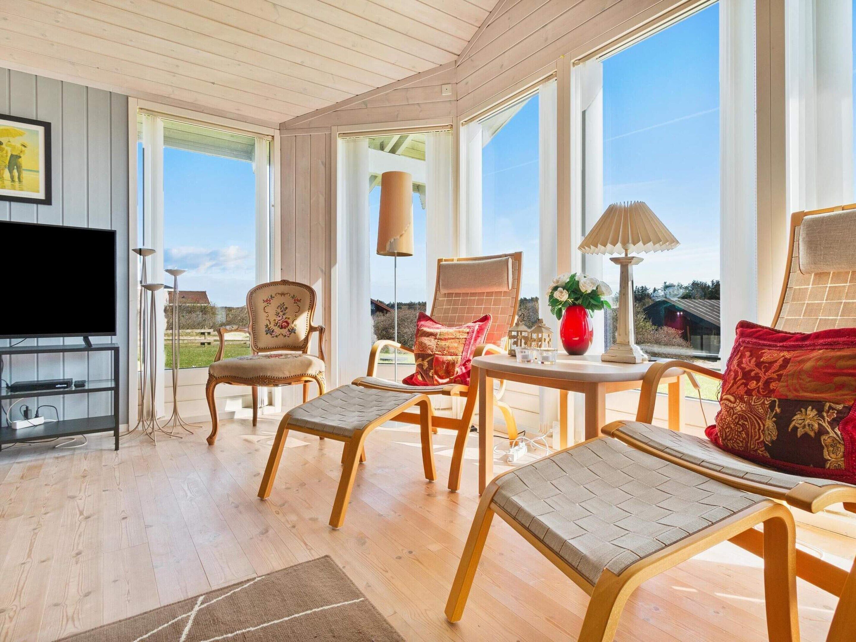 Una habitación iluminada por el sol con suelos de madera, un televisor, un sofá, dos sillones, una mesa con una lámpara y grandes ventanas que ofrecen vistas al exterior.