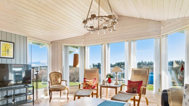 A spacious living room with a wooden ceiling, large windows, a chandelier, and a television.