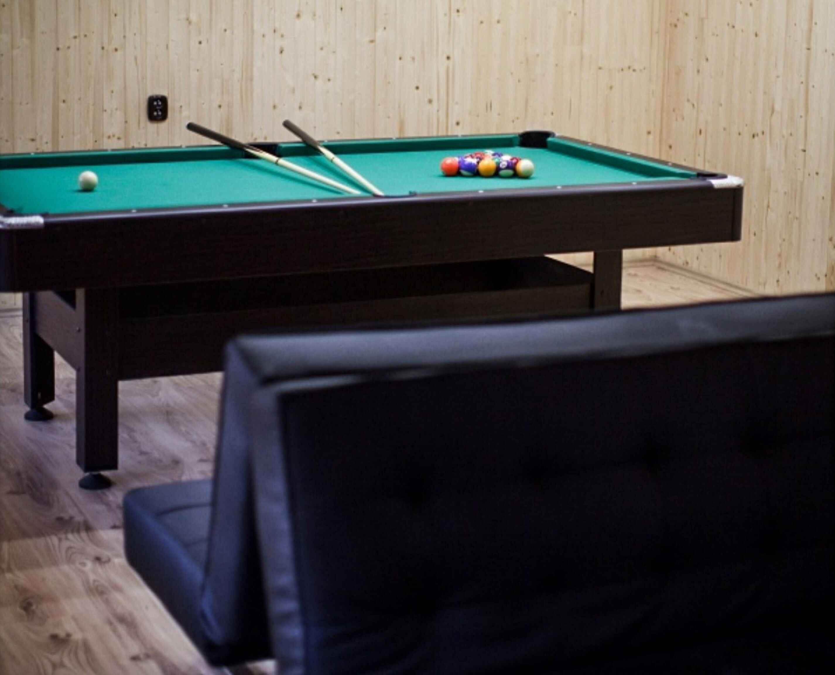 A pool table with balls and cues, a wooden wall, and a black chair in the foreground.