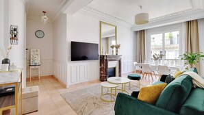 A spacious living room with a green sofa, a flat-screen TV, a fireplace, and a dining area with a table and chairs. - Modern Apartment - 2bd/4p - Louvre/marais (Paris)