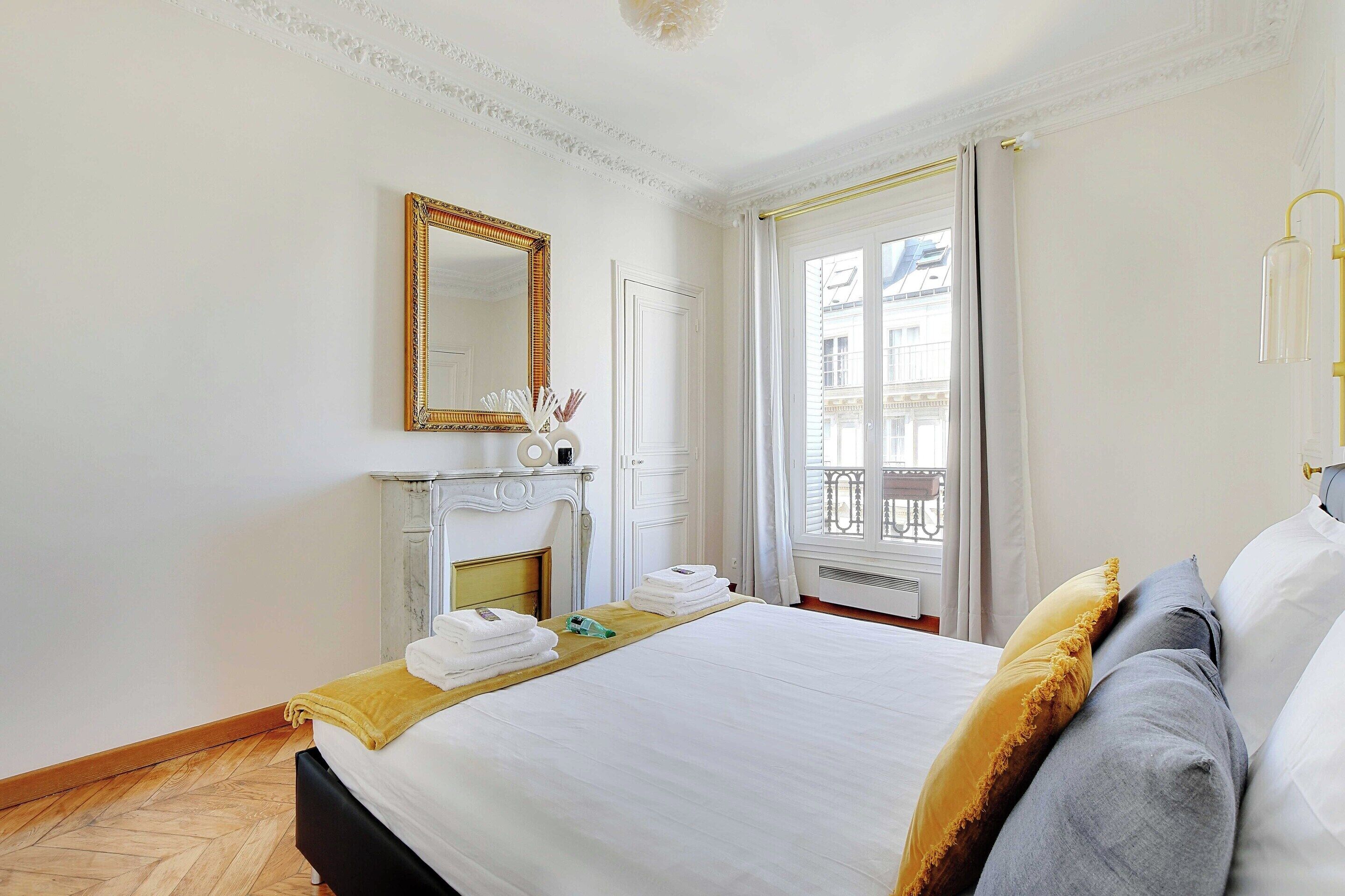 A hotel room with a large bed, a fireplace, a mirror, and a view of the city through the window.