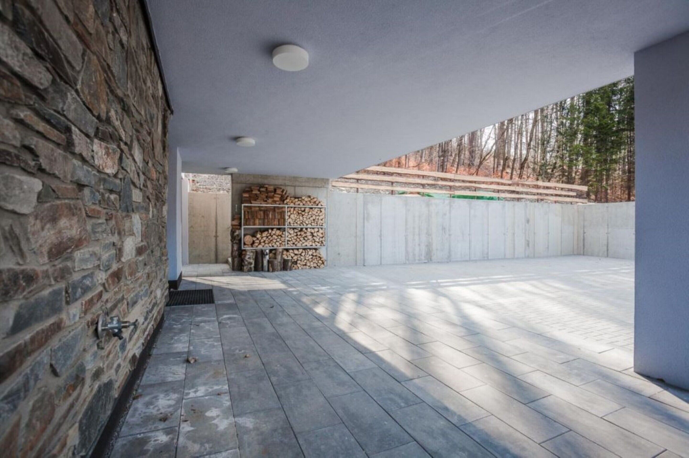 A covered outdoor area with a stone wall, a firewood stack, and a view of trees.
