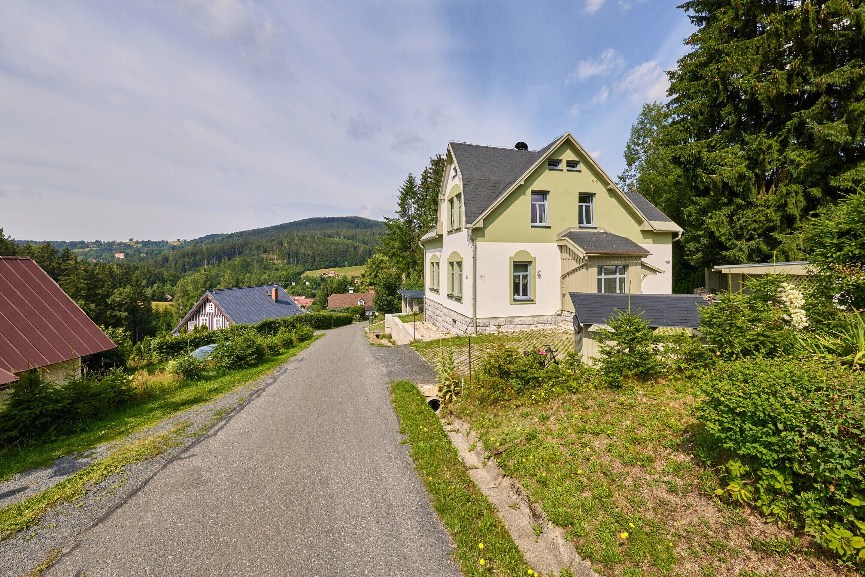 Eine ländliche Straße führt zu einem großen, hellen Haus mit steilem Dach, umgeben von viel Grün und einer fernen Bergkette.