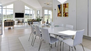 Un espace de repas moderne avec une table et des chaises blanches, de grandes fenêtres et un téléviseur à écran plat en arrière-plan.
