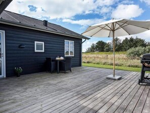 A wooden deck with a barbecue grill and an umbrella, a dark-colored house with two windows, and a grassy field in the background. - 5 Star Holiday Home in Lokken (Lokken)