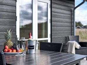 A table with a fruit bowl, champagne bottle, and glasses, set on a deck with a view of a house. - 5 Star Holiday Home in Lokken (Lokken)