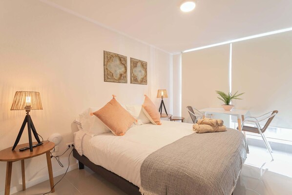A bedroom with a bed, bedside lamps, a table, a chair, and a potted plant.