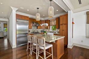A modern kitchen with wooden cabinets, a stainless steel refrigerator, a large island with bar stools, and pendant lighting. - Hale Akea by Alekona Kauai: Charming 3br Cottage w AC, Ocean Views, Spa, large yard (Koloa)