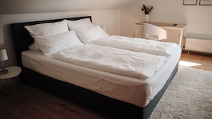 Uma cama de casal bem arrumada com roupa de cama branca num quarto moderno com soalho de madeira, uma mesa de cabeceira com uma lâmpada e uma pequena mesa de cabeceira com uma planta.
