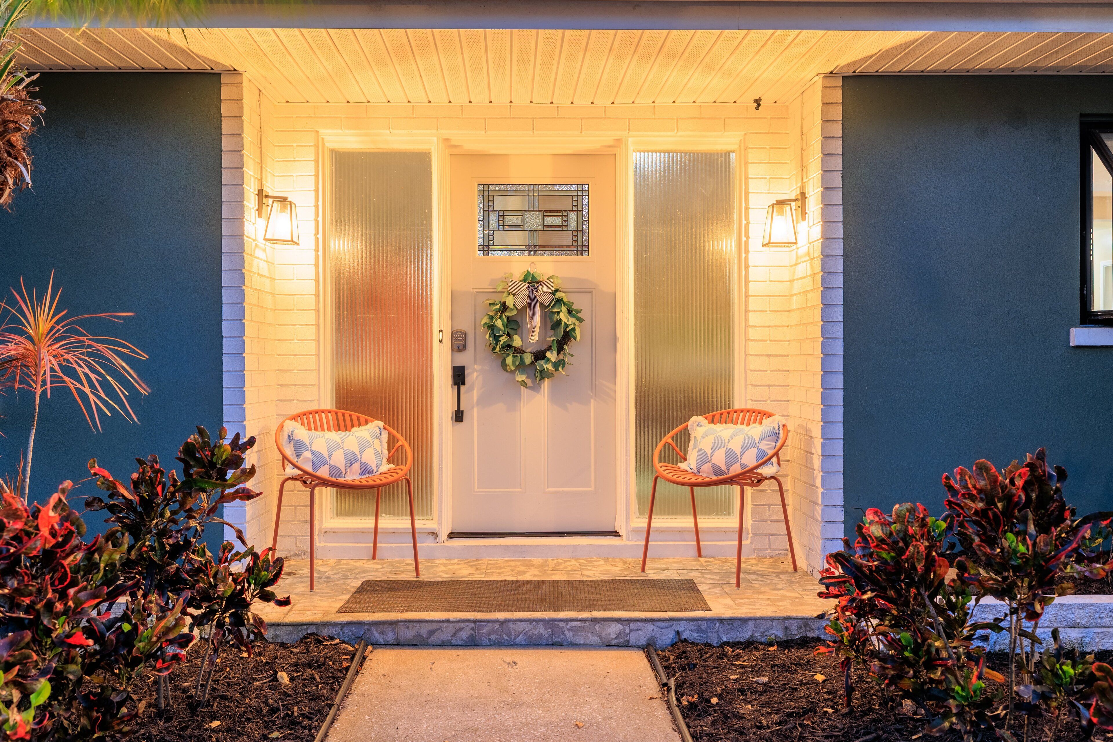 A house entrance with a white door, two wicker chairs, a wreath, and a small window above the door.