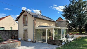 Une maison avec une véranda en verre, une allée gravillonnée et un jardin agrémenté de plantes et d’un arbre.