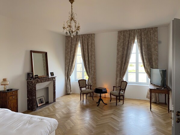 Une pièce spacieuse avec un grand lit, une cheminée, deux fauteuils, un téléviseur et un lustre.