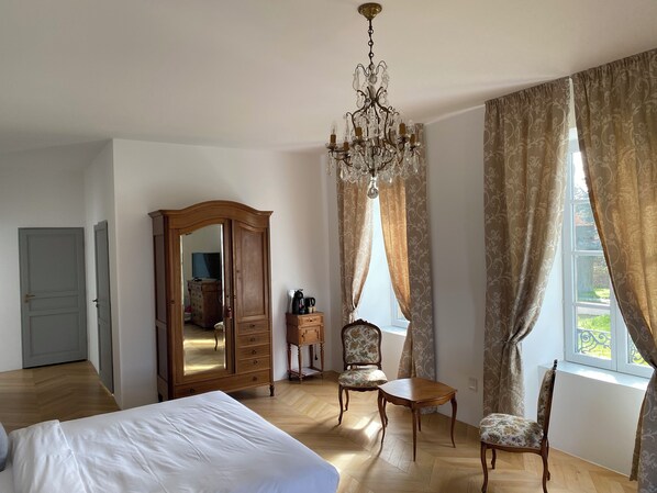 A hotel room with a large bed, a wooden wardrobe, a chandelier, a dining table, two chairs, and a window with curtains.