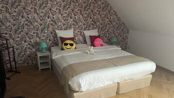 A bedroom with a large bed, a wooden floor, a floral wallpaper, and a small nightstand with a lamp.