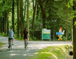 Duas pessoas andando de bicicleta em um caminho pavimentado, rodeado por árvores. Há uma placa para 'Ruwinkel' e um sinal de estacionamento 'P' com um mascote de pato.