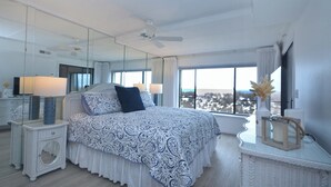 A bedroom with a large bed, a ceiling fan, a window with a view, and a bedside table with a lamp.