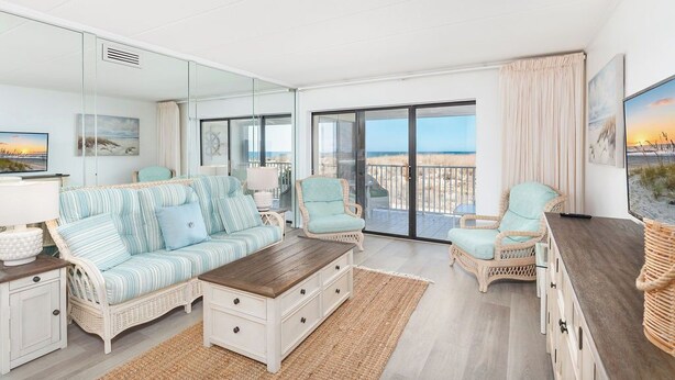 A spacious living room with a large window offering a view of the beach, a striped sofa, wicker chairs, and a wooden coffee table.