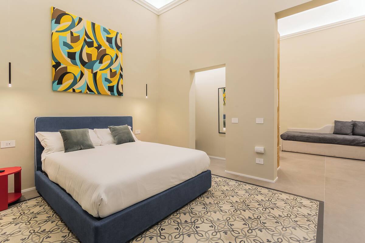 Un dormitorio moderno con una cama grande, una mesita de noche roja, un tapete estampado y un cuadro abstracto colorido en la pared.