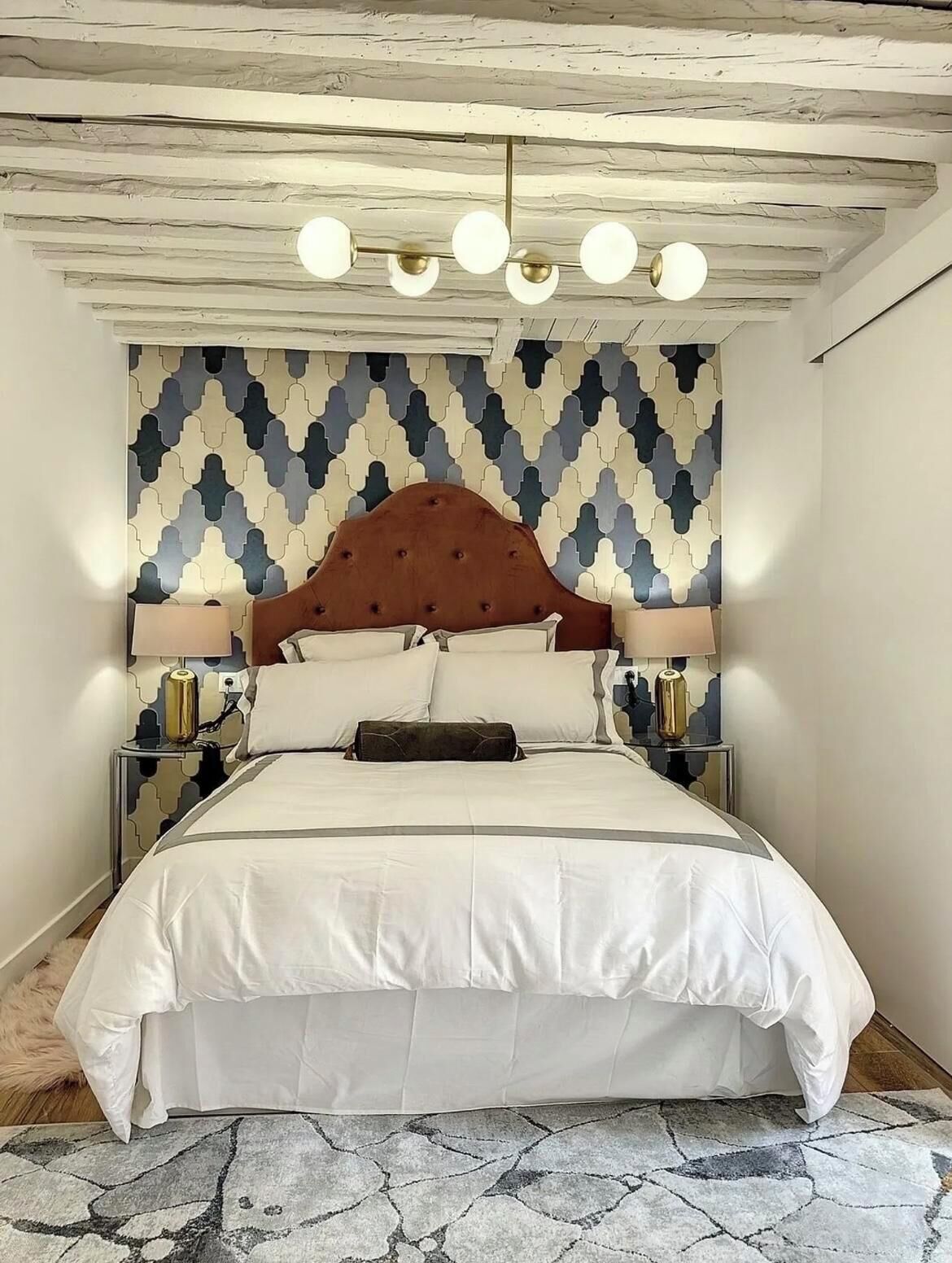 Ein ordentlich bezogenes Bett mit Holz-Kopfstück, einer Wand mit Muster und einer Decke mit freiliegenden Balken und Lampen.