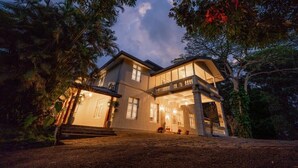 Une grande maison à deux étages, dotée d’un balcon et d’un porche, entourée d’arbres et bordée d’une allée en gravier.