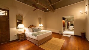 Une chambre avec un plancher en bois, un lit avec une literie blanche, deux lampes de chevet, un ventilateur de plafond, un miroir et une vue sur une autre pièce.