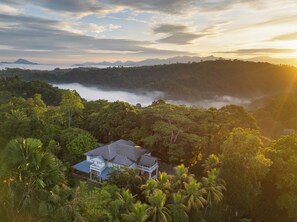 A house surrounded by dense tropical vegetation and a misty mountain range in the background.