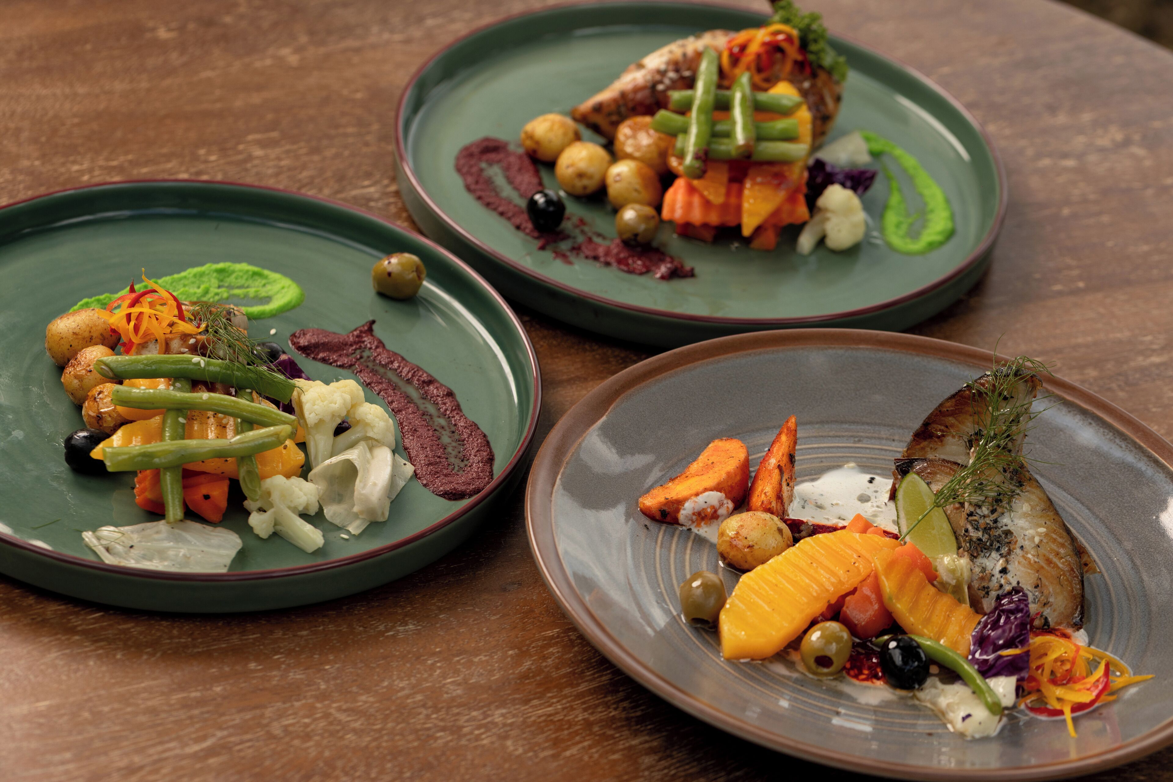 Trois assiettes de nourriture, composées de divers légumes et de poisson, servies sur une table en bois.