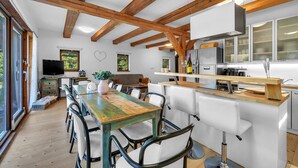 A modern kitchen with wooden beams, a dining area with a wooden table and chairs, and a large window overlooking greenery.