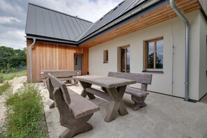 A modern outdoor dining area with wooden benches and a stone table, surrounded by a white building with large windows and a sloped roof. - Child-friendly Villa in Bohemia (Korenov)