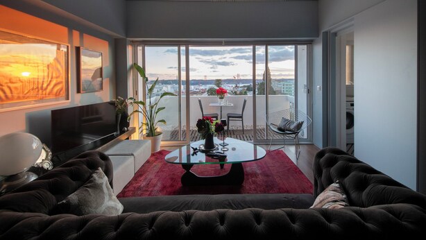 A modern living room with a large sofa, a glass coffee table, and a red rug. There is a balcony with a view of the city and a table with chairs.