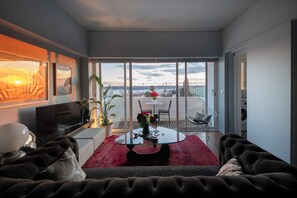 A modern living room with a large sofa, a glass coffee table, and a red rug. There is a balcony with a view of the city and a table with chairs. - Lisbon river view design penthouse (Lisboa)