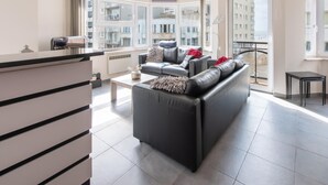 A modern living room with a black leather sofa, a coffee table, and a view of a cityscape through large windows. - Beautiful Apartment With Lateral sea View (Ostend)