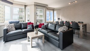A modern living room with a black leather sofa, a wooden coffee table, and a dining area with a table and chairs. - Beautiful Apartment With Lateral sea View (Ostend)