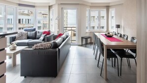 A modern living room with a sofa, a dining table, and chairs. There are large windows with a view of buildings outside. - Beautiful Apartment With Lateral sea View (Ostend)