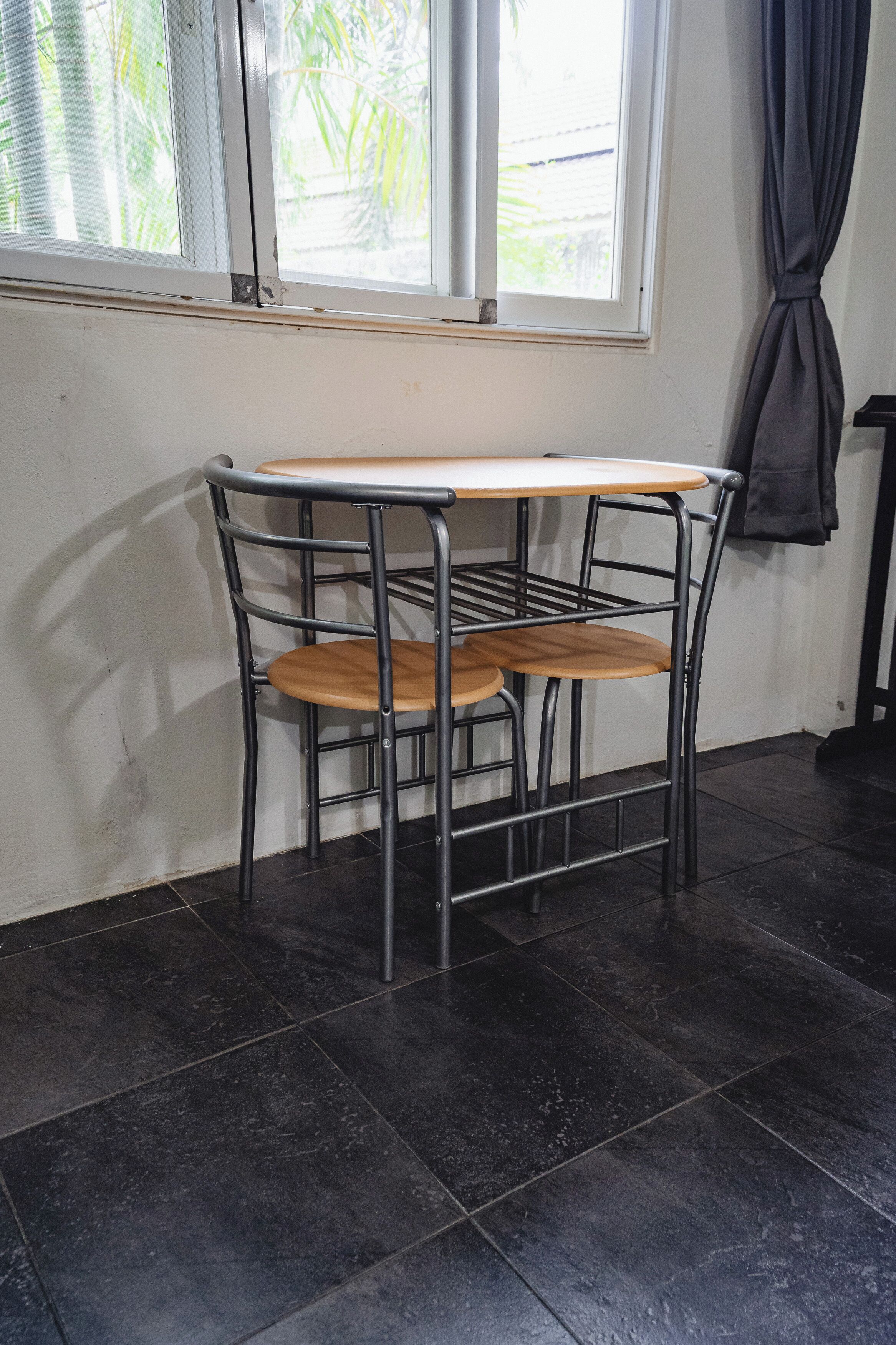 A small, metal-framed table with a wooden top and two chairs, positioned next to a window with a view of greenery outside.