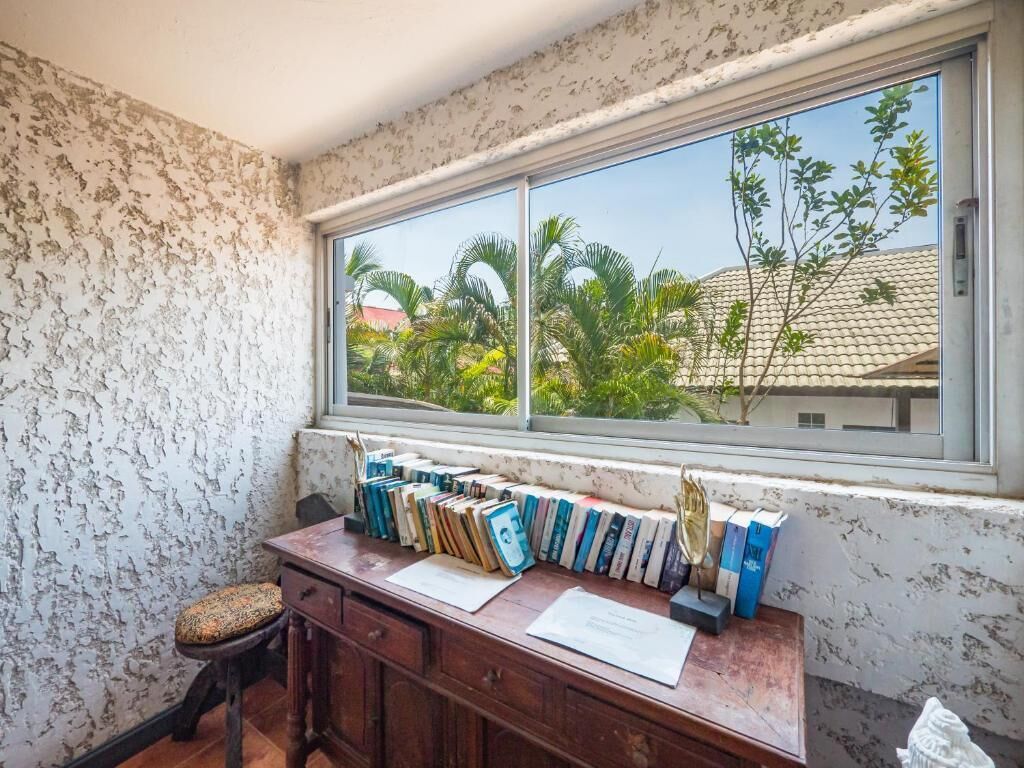 A room with a window overlooking a garden, a wooden desk with books and papers, and a small stool.