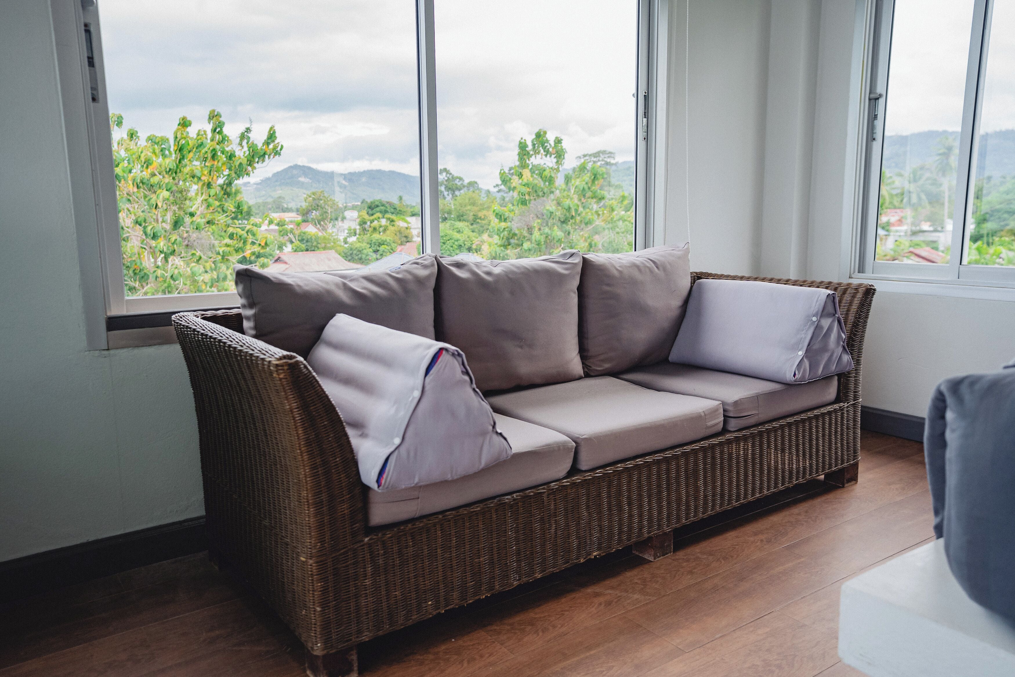 Un sofá de mimbre con cojines grises en una habitación con ventanas amplias que dan a vistas de vegetación y colinas.