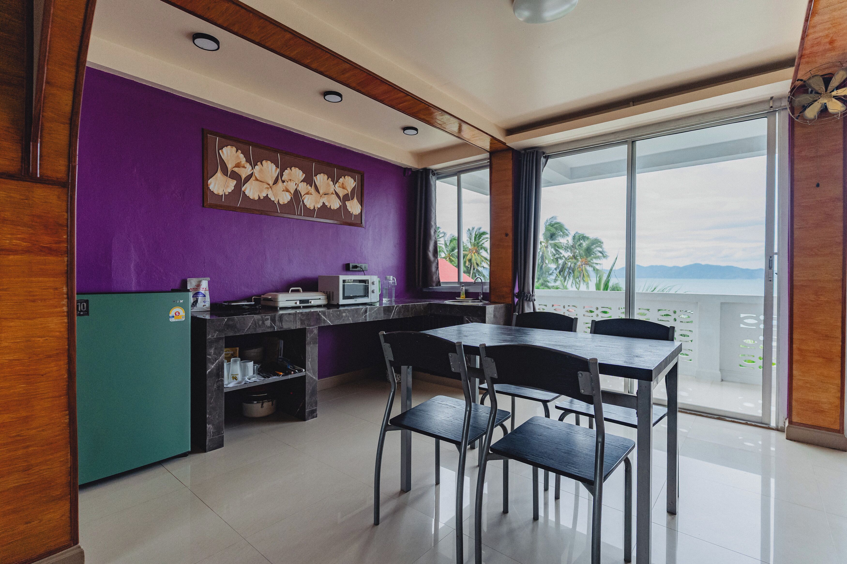 Una cocina moderna con una pared morada, una zona de comedor con cuatro sillas y vistas a palmeras a través de amplias ventanas.