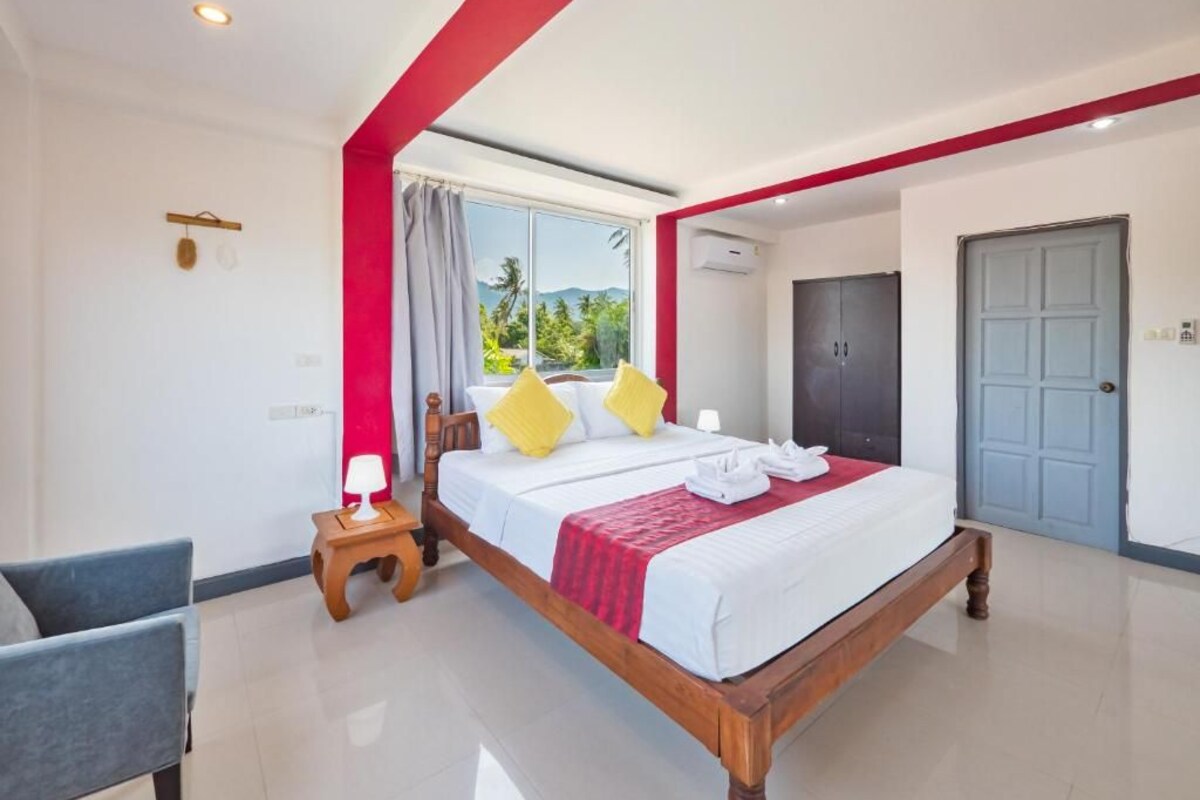 A hotel room with a wooden bed, a small bedside table, a chair, a wardrobe, and a window with a view of greenery.